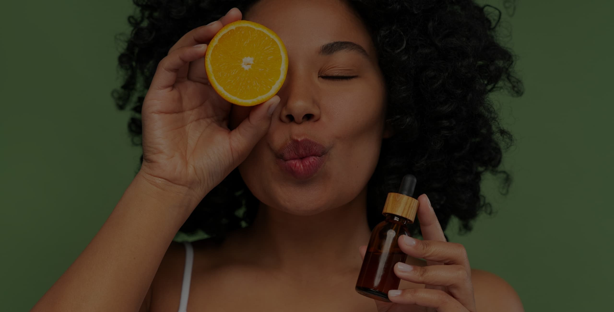 Woman holding a serum bottle and orange slice over one eye